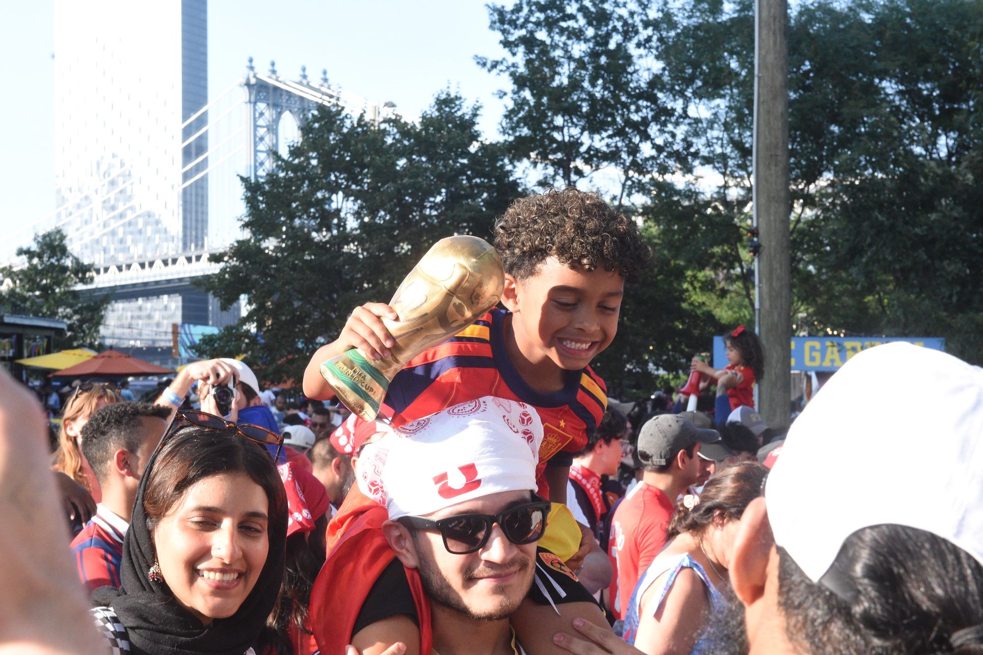 A crowd of people celebrating, with a young boy holding a trophy and smiling in the foreground. The Manhattan Bridge can be seen in the background.