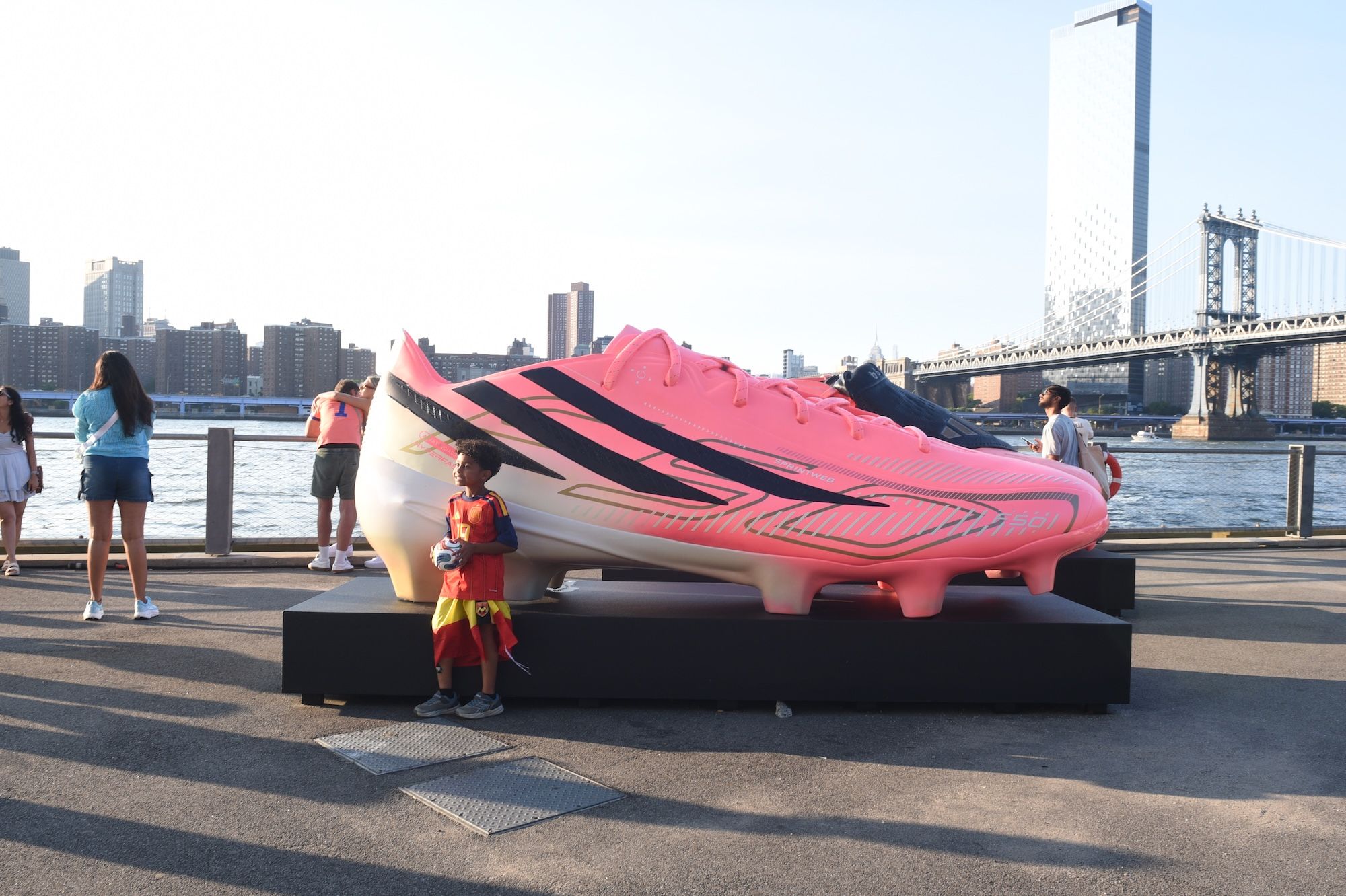Large pink and black soccer cleat sculpture on a waterfront promenade with a cityscape in the background and people standing nearby.