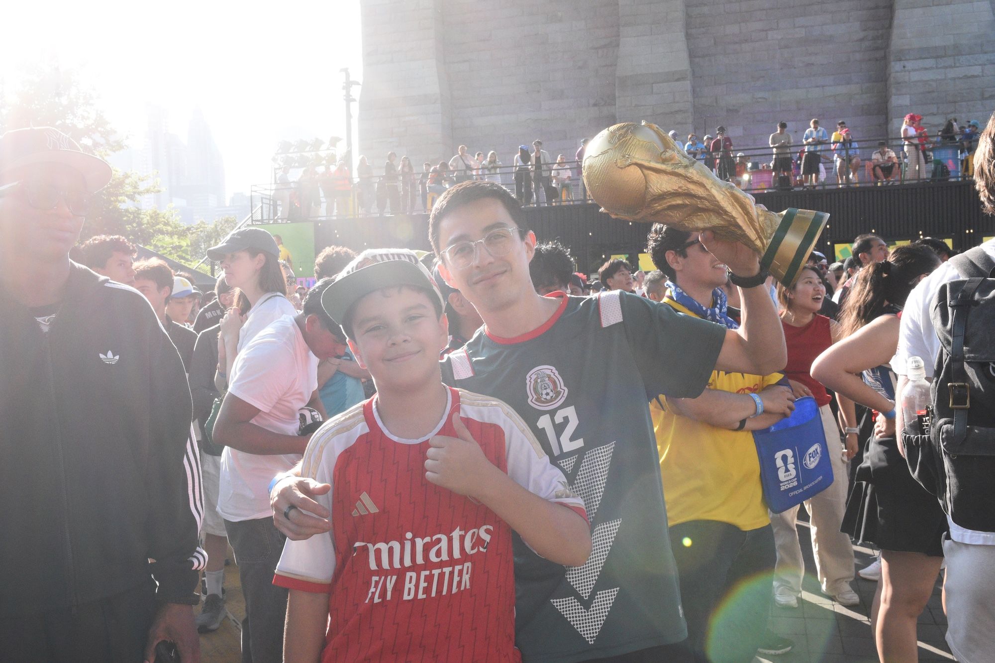 A group of people, including two young boys, posing for a photo at a crowded outdoor event. One boy is wearing a red soccer jersey with the "Emirates" logo, and the other is holding a large trophy.
