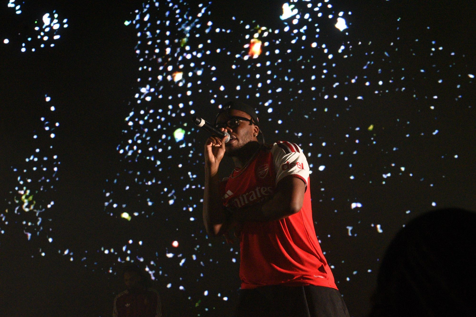 A performer wearing a red jersey with the word "Emirates" on it singing on stage against a backdrop of colorful fireworks.