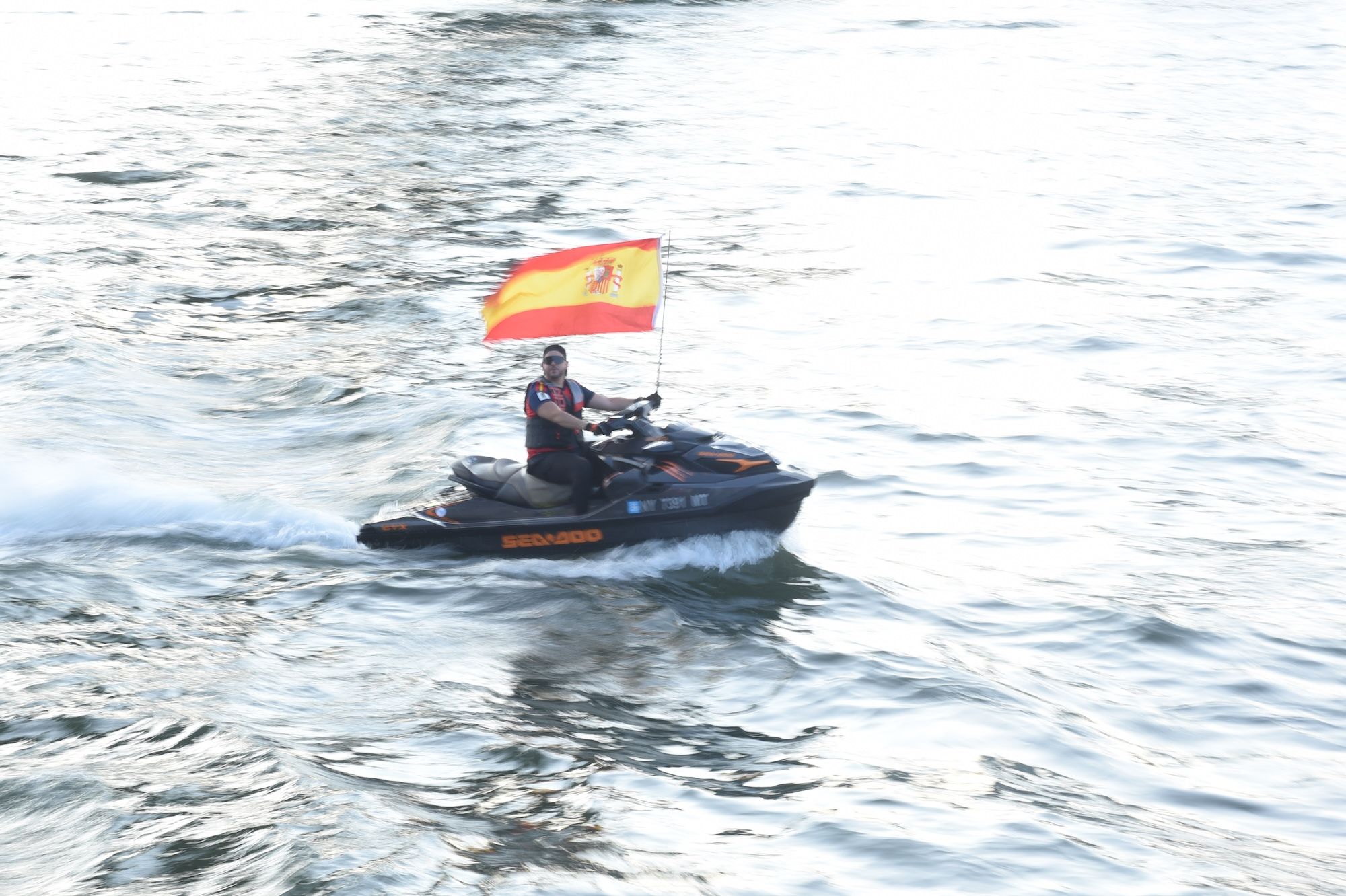 A person riding a Seadoo jet ski on the water, waving a Spanish flag.