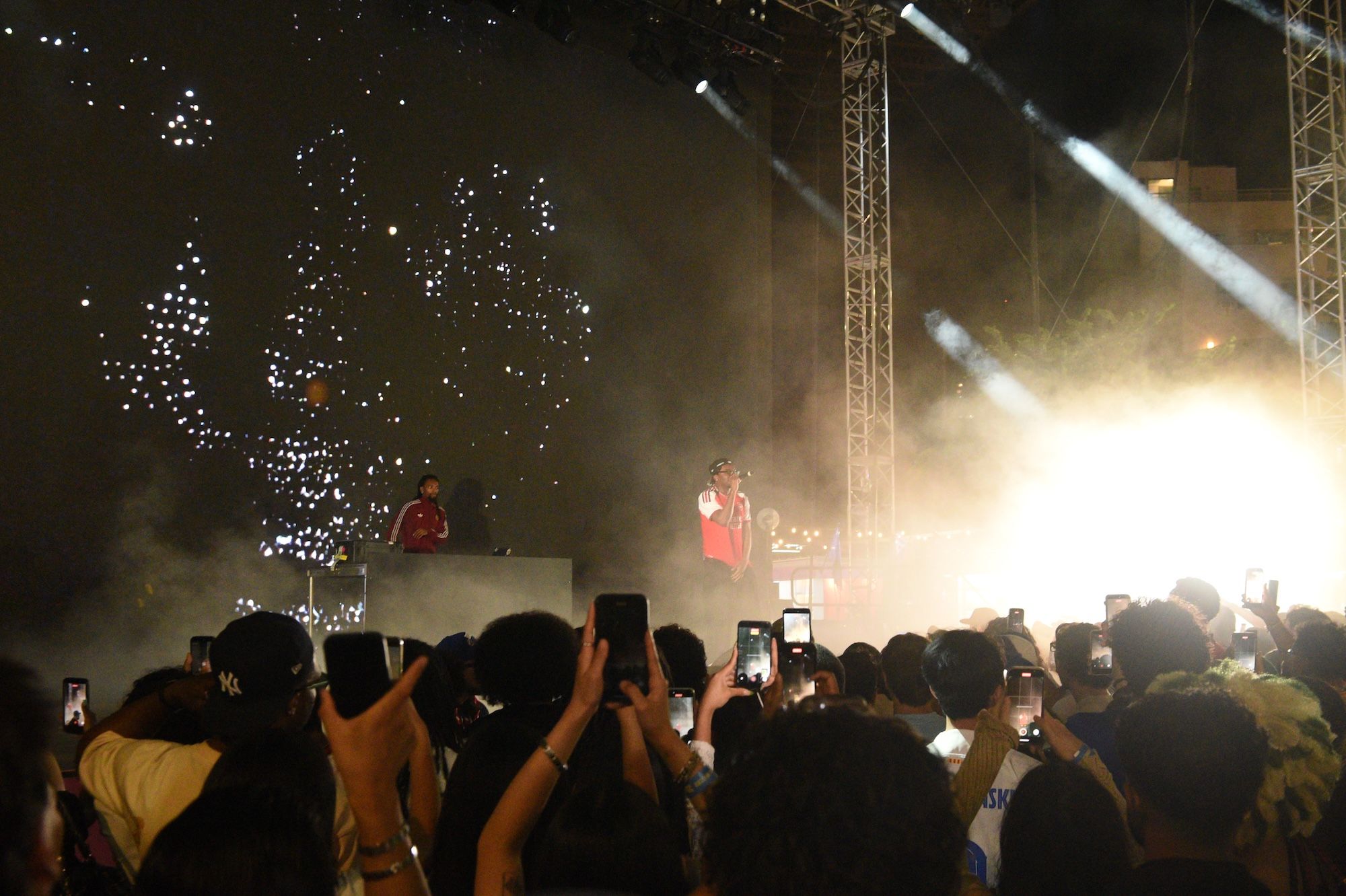 Nighttime concert scene with crowd holding up phones, performers on stage, and fireworks display in the sky.