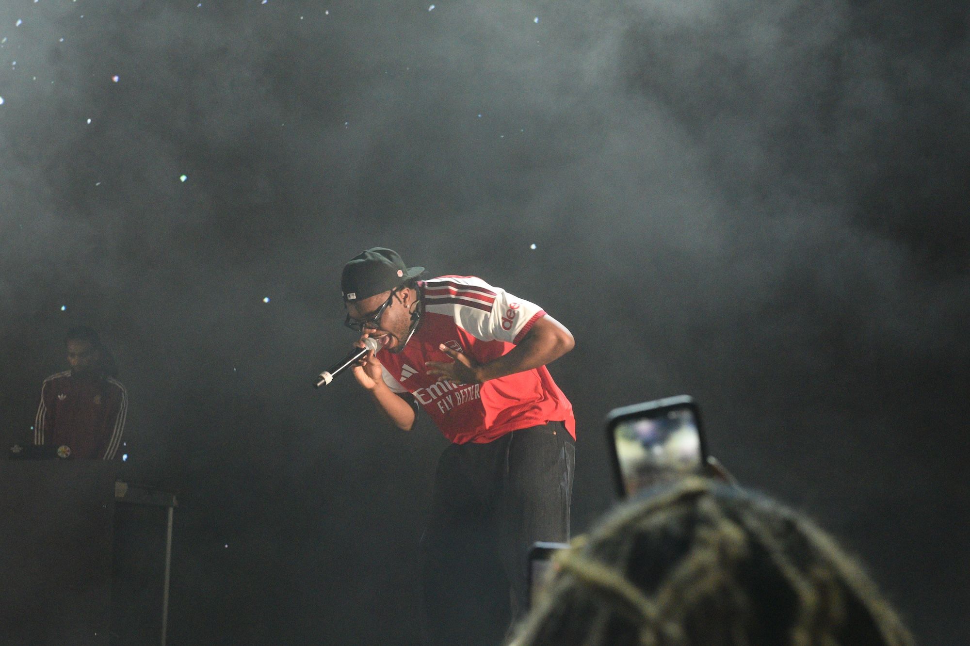 A person wearing a red jersey and cap performing on stage in a dark, smoky environment with stars in the background.