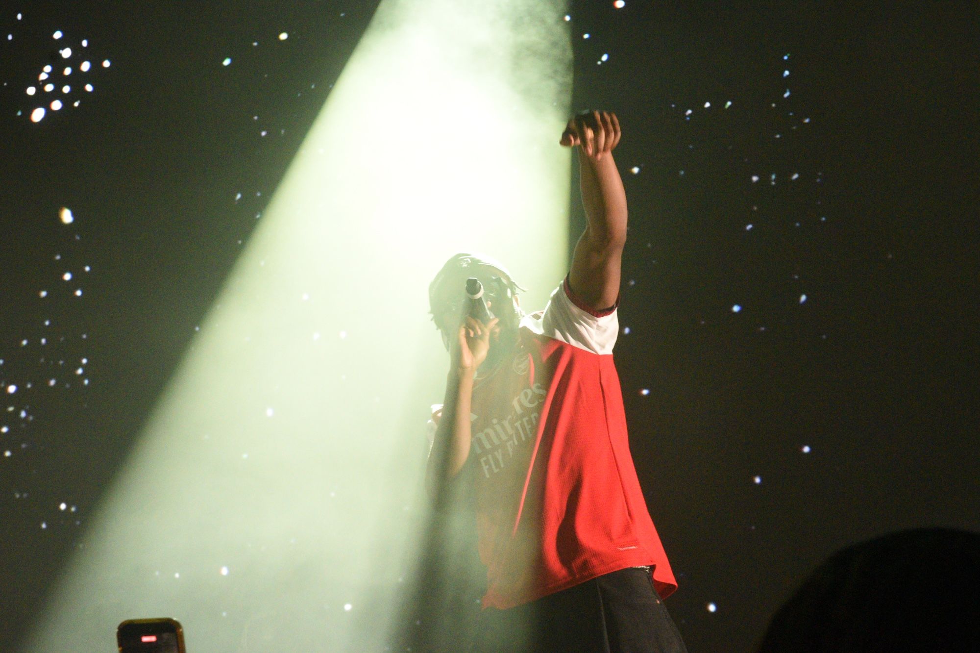 A person in a red shirt raising their arms on a stage with spotlights and stars in the background.