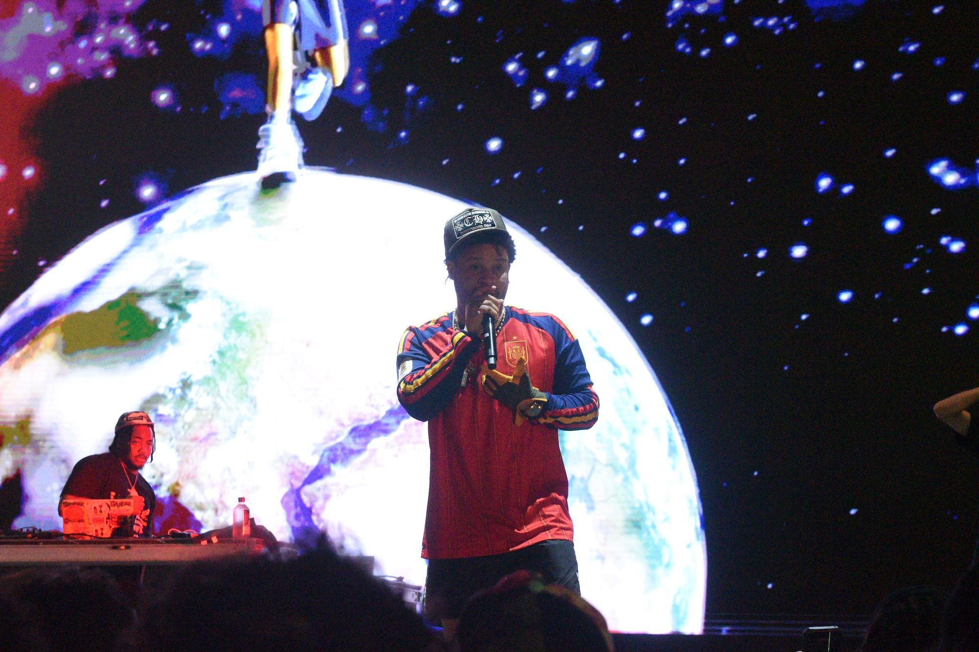 A performer on stage in a red and blue outfit holding a microphone, with a large glowing planet-like sphere behind them and stars in the background.