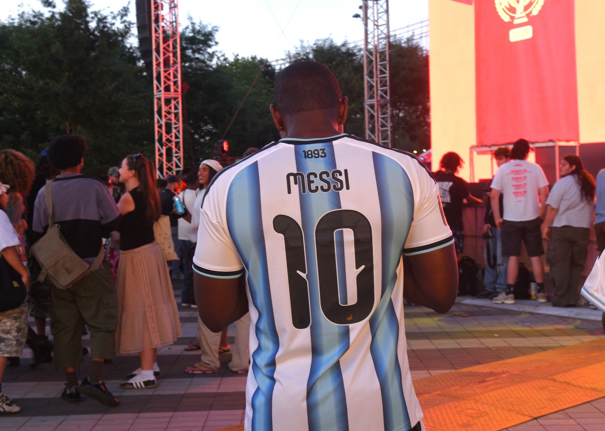 A person wearing a white and blue soccer jersey with the number "10" and the name "Messi" on the back, standing in a crowd at an event with red and white banners in the background.