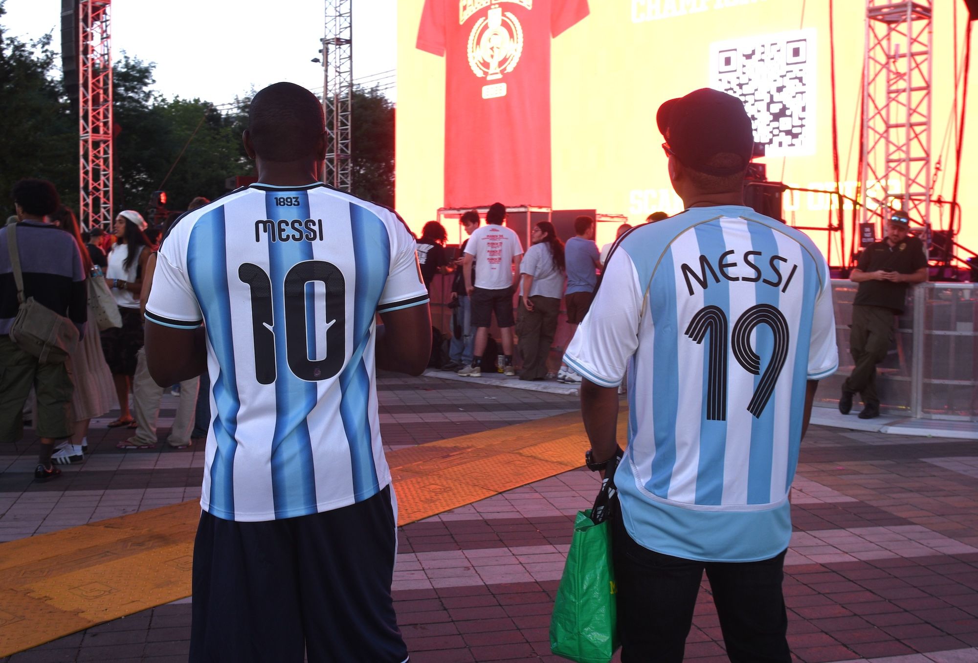 Two people wearing Argentina soccer jerseys with the name "Messi" on the back, standing in front of a large stage or event venue.