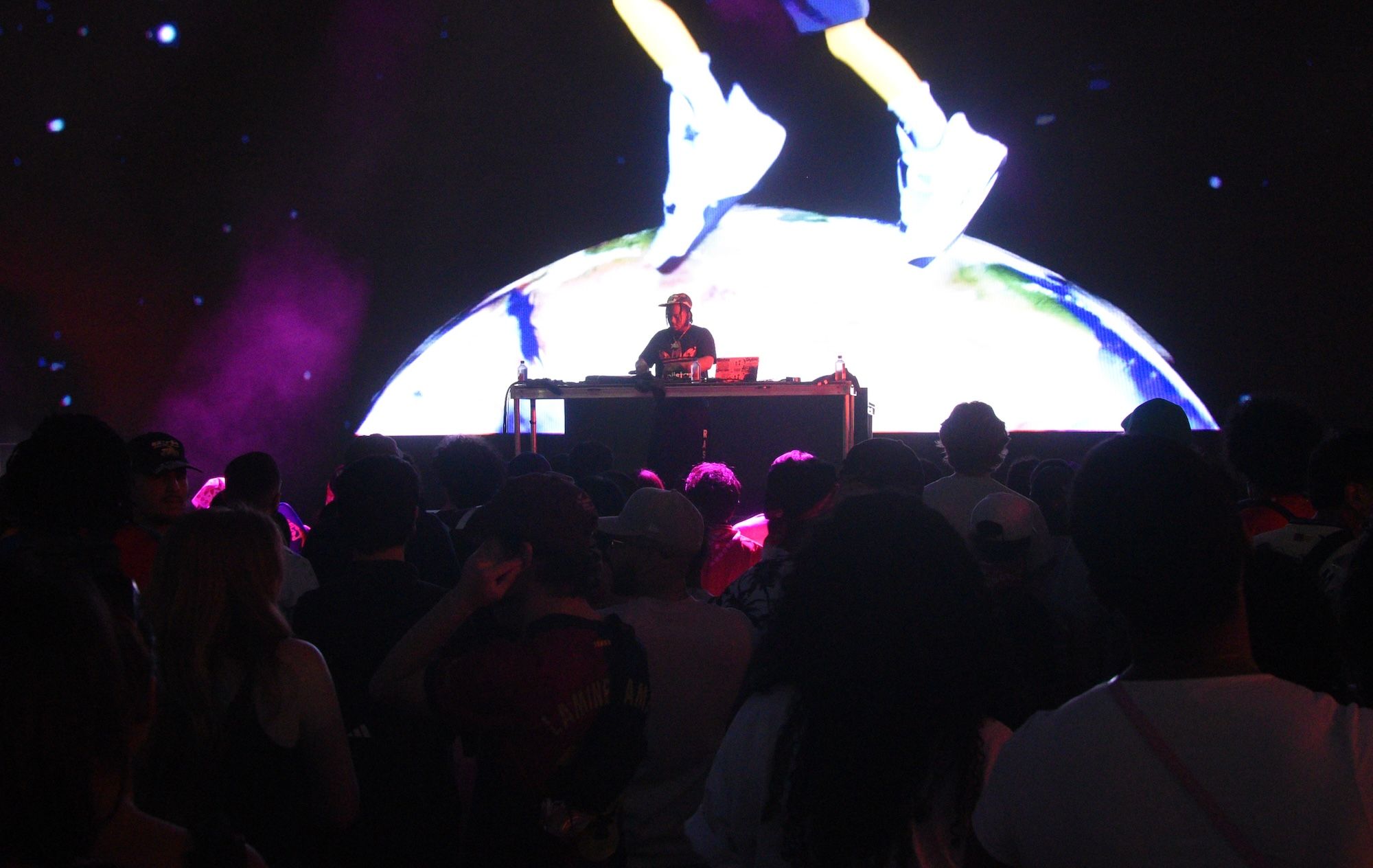A DJ performing on stage in front of a large crowd, with a colorful, illuminated globe-like projection behind them.