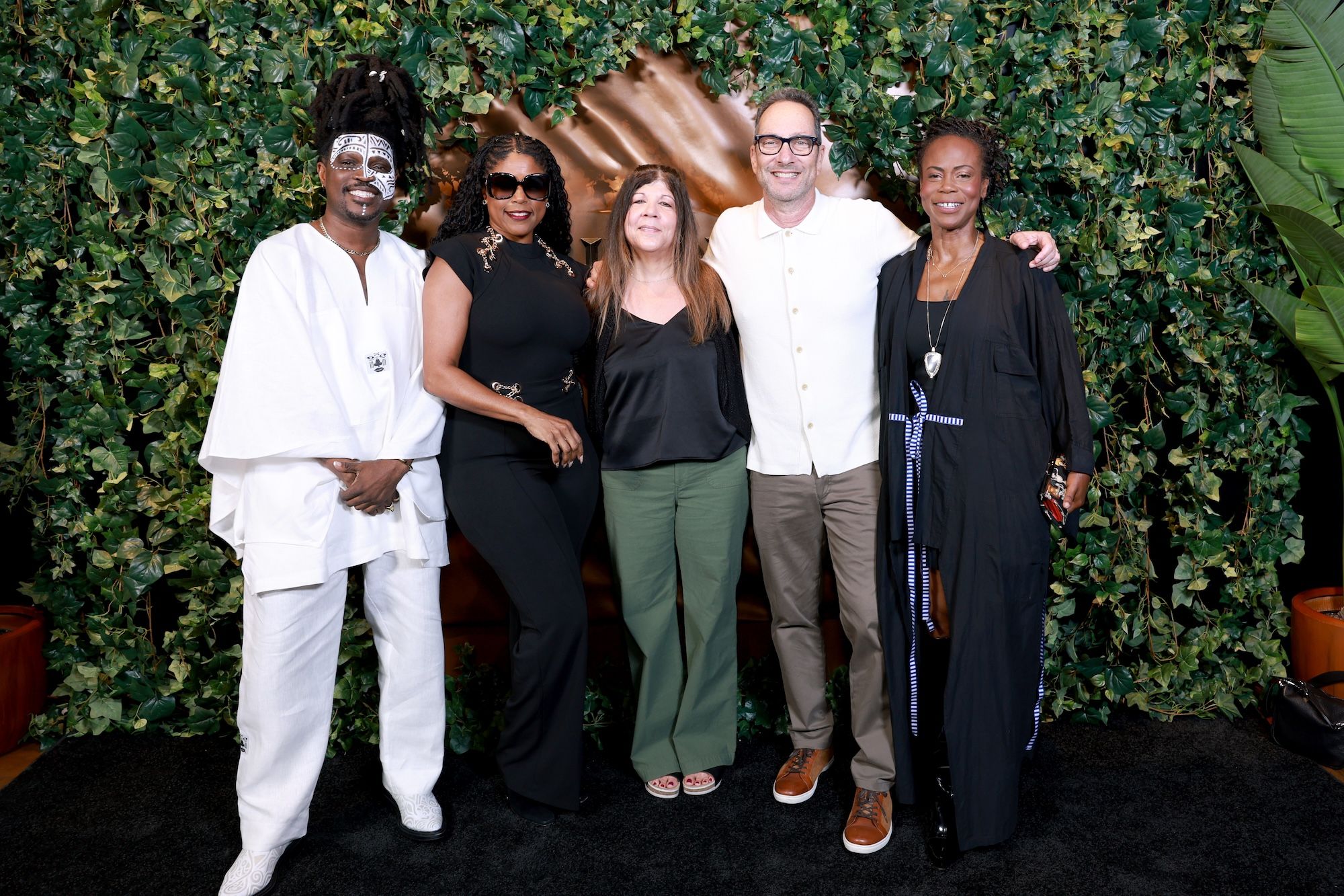 A group of five people, three women and two men, standing together in front of a lush green backdrop. They are dressed in formal attire, with the women wearing black and the men wearing white and tan.