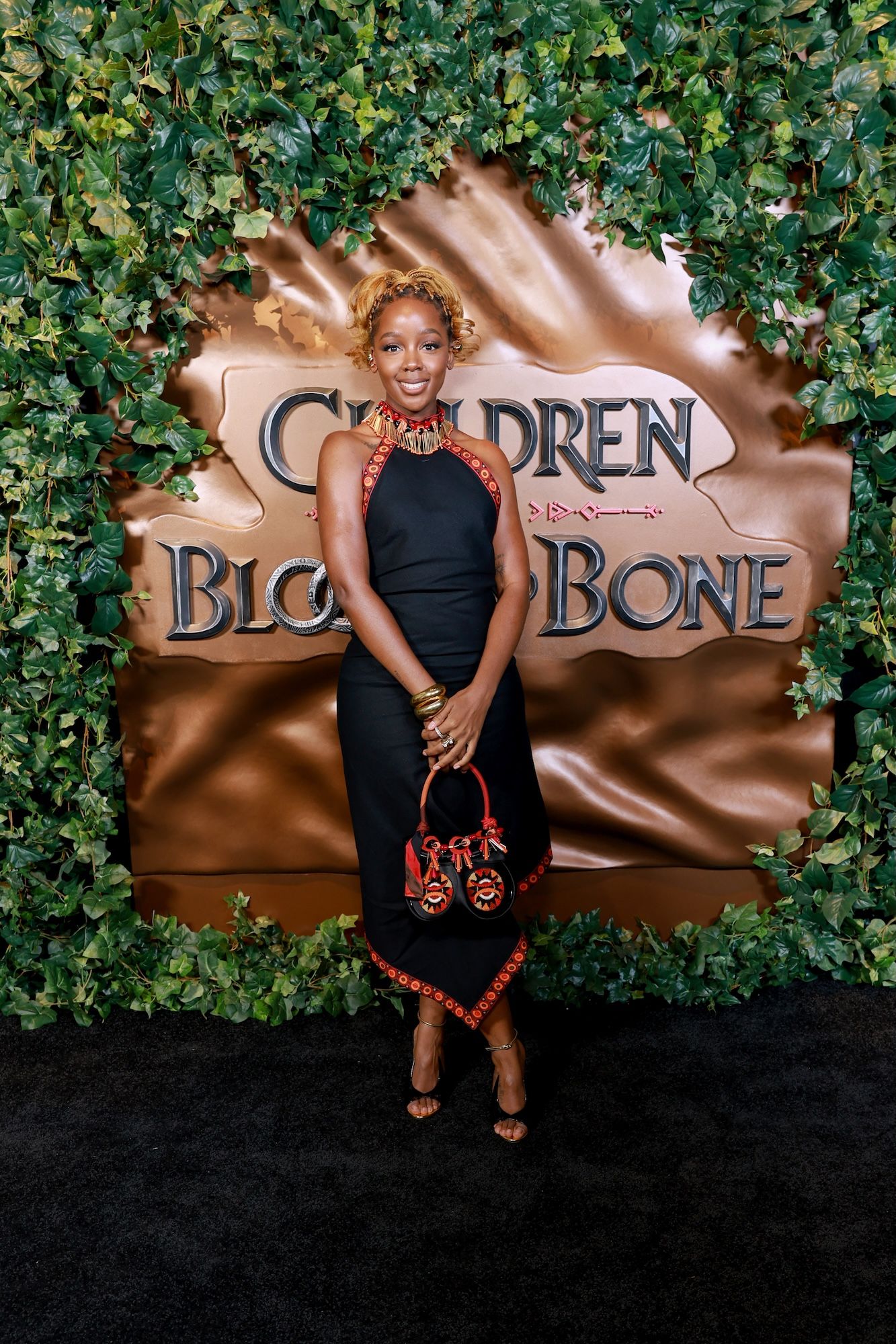 A woman wearing a black dress and colorful accessories stands in front of a large sign that says "Children of Blood and Bone".