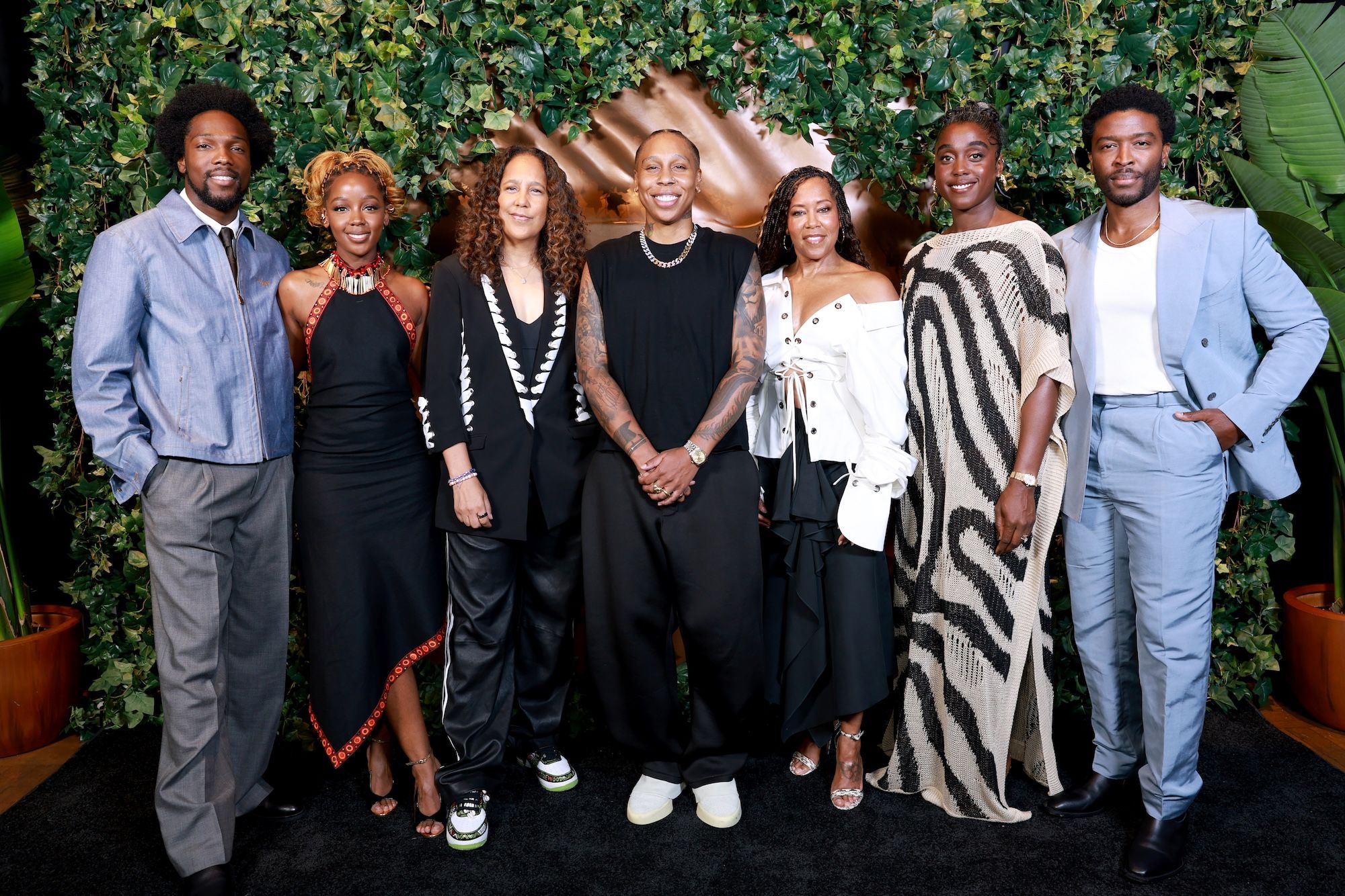 A group of 7 Black individuals, including actors Tosin Cole, Thuso Mbedu, Lena Waithe, Regina King, Lashana Lynch, and director Gina Prince-Bythewood, posing together in front of a lush green backdrop.