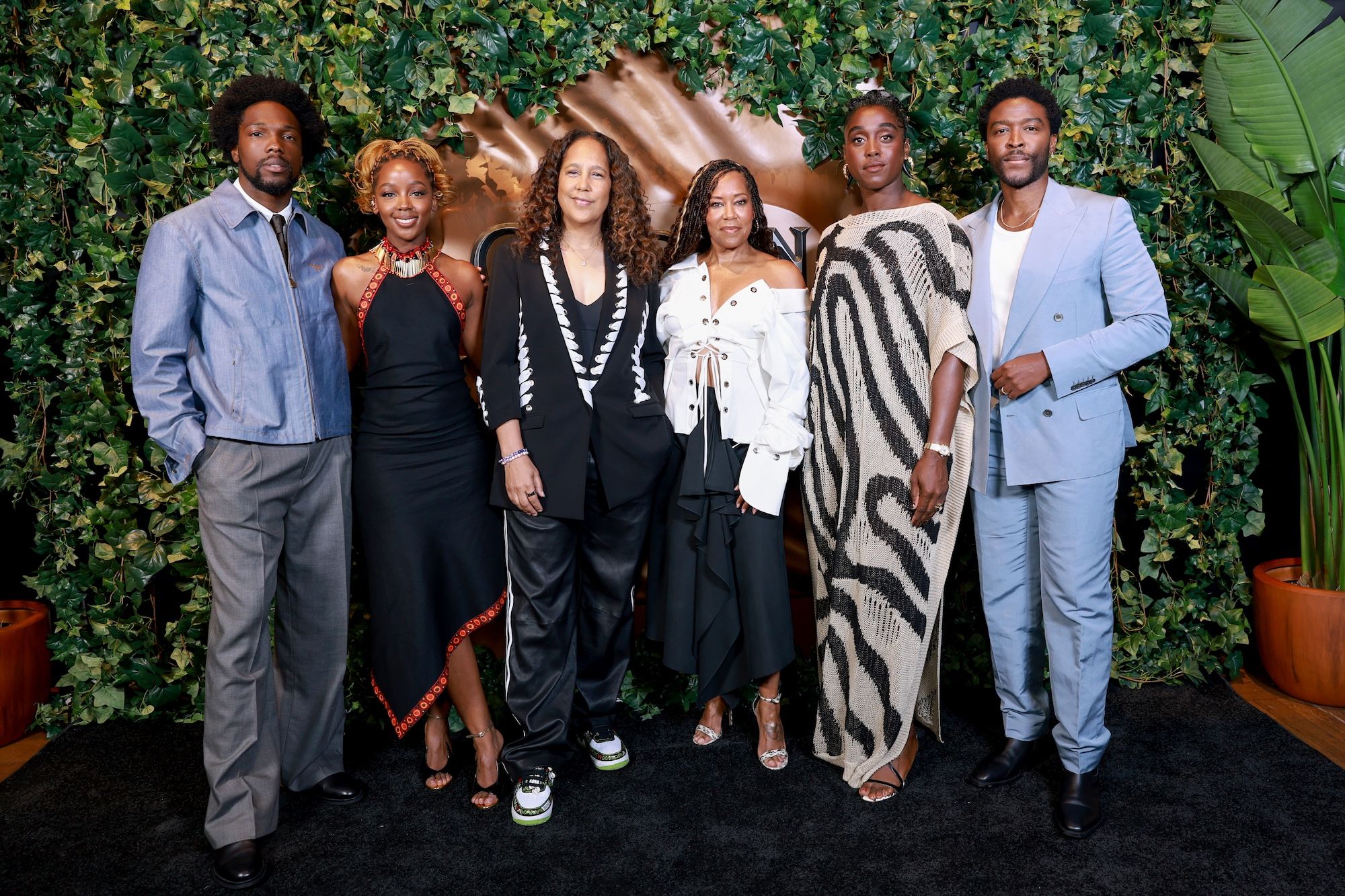 A group of six people, including actors Tosin Cole, Thuso Mbedu, Regina King, Lashana Lynch, and Zackary Momoh, posing together in front of a lush green backdrop.
