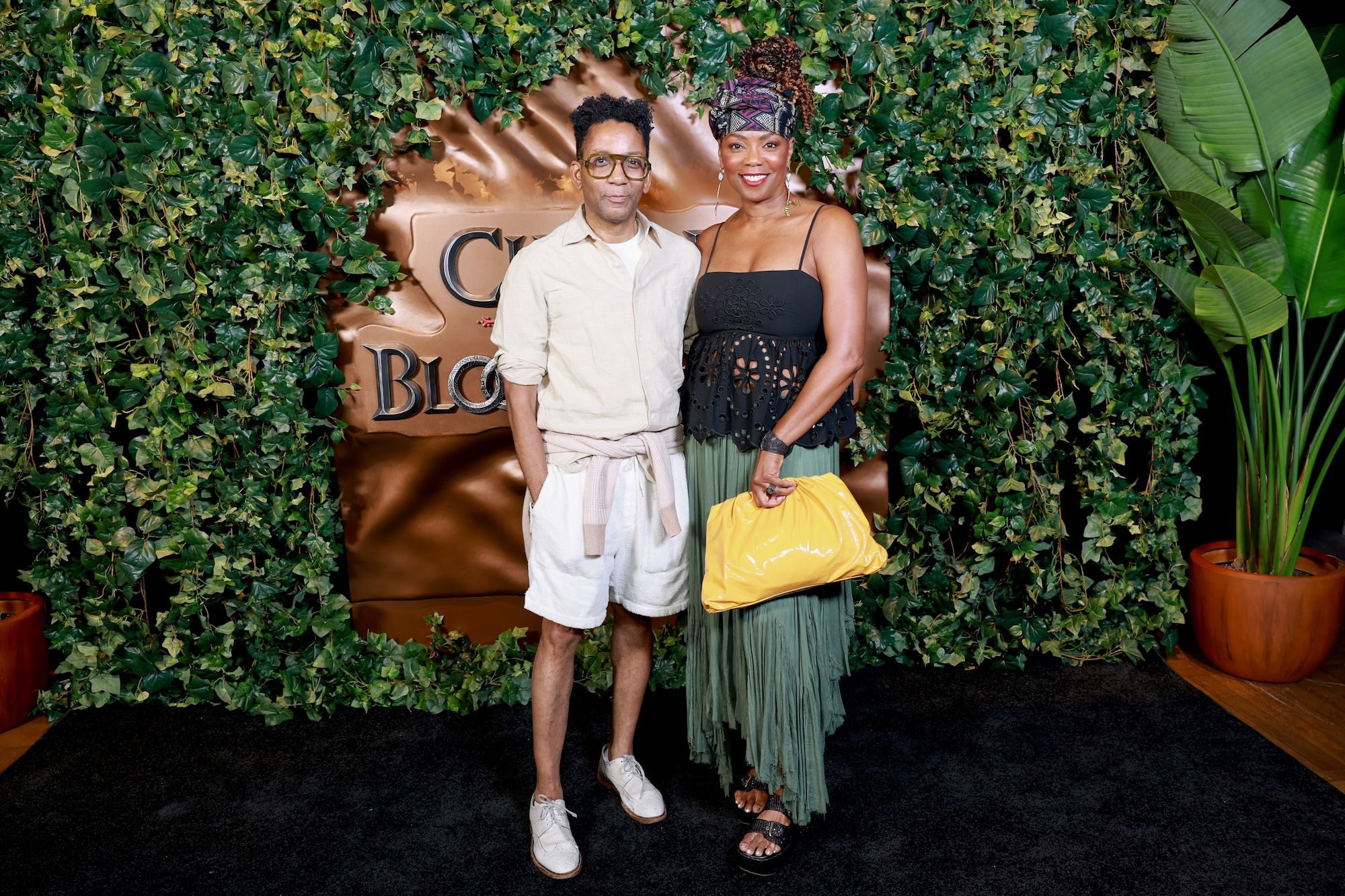 Two people posing in front of a green foliage backdrop with a large "Coco Bongo" sign. The person on the left is wearing a light-colored outfit, and the person on the right is wearing a black dress with a colorful headpiece.