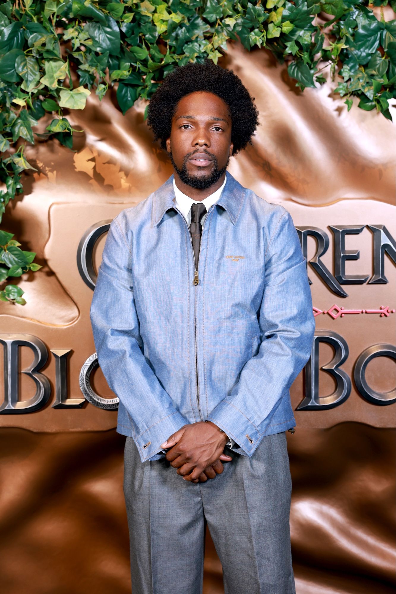 A man with a beard and afro hairstyle wearing a denim jacket and tie stands in front of a backdrop with the text "CHILDREN OF BLOOD AND BONE".