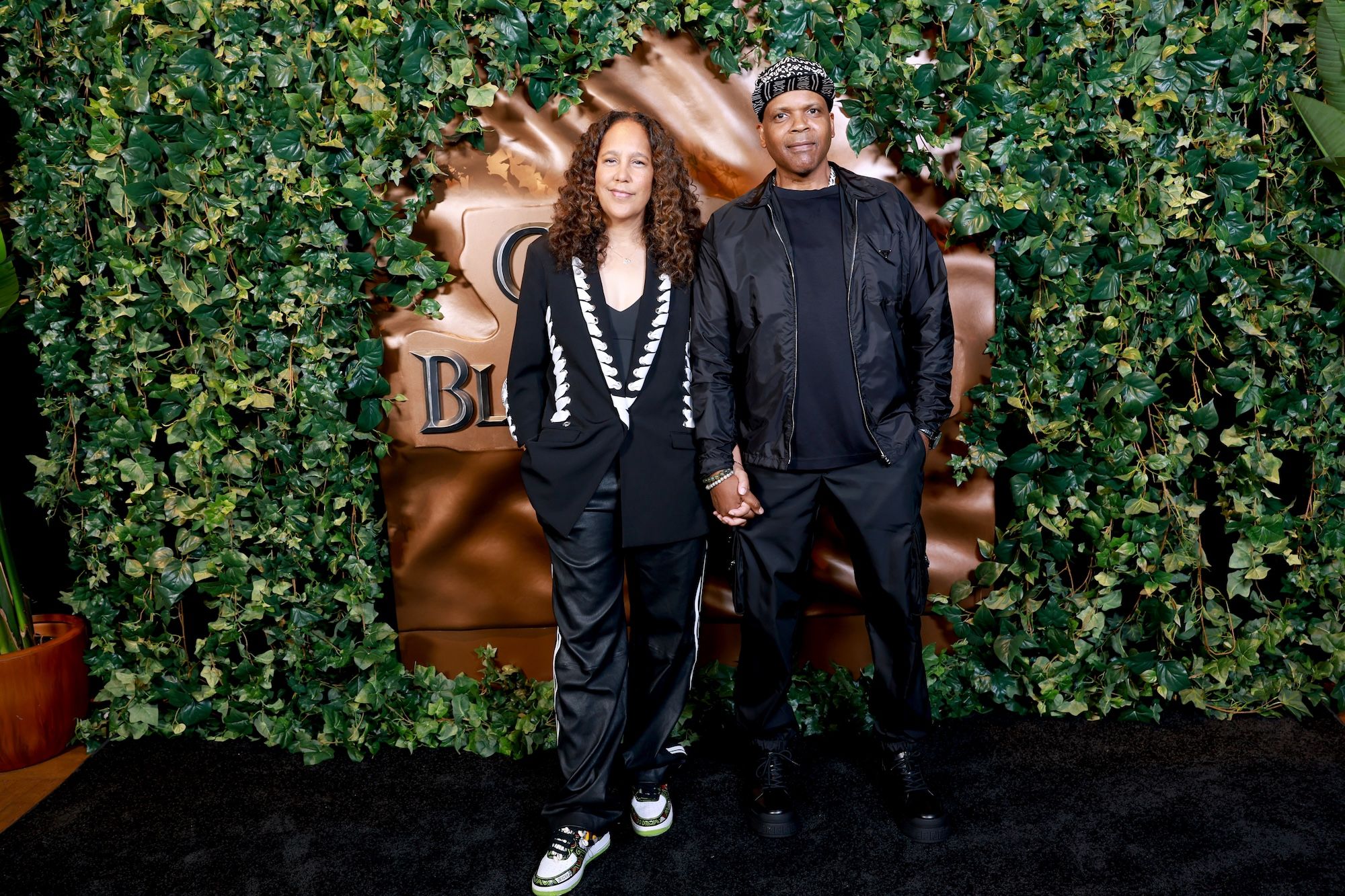Two people, a woman with curly hair and a man, standing together in front of a large "BLOOM" sign surrounded by greenery.