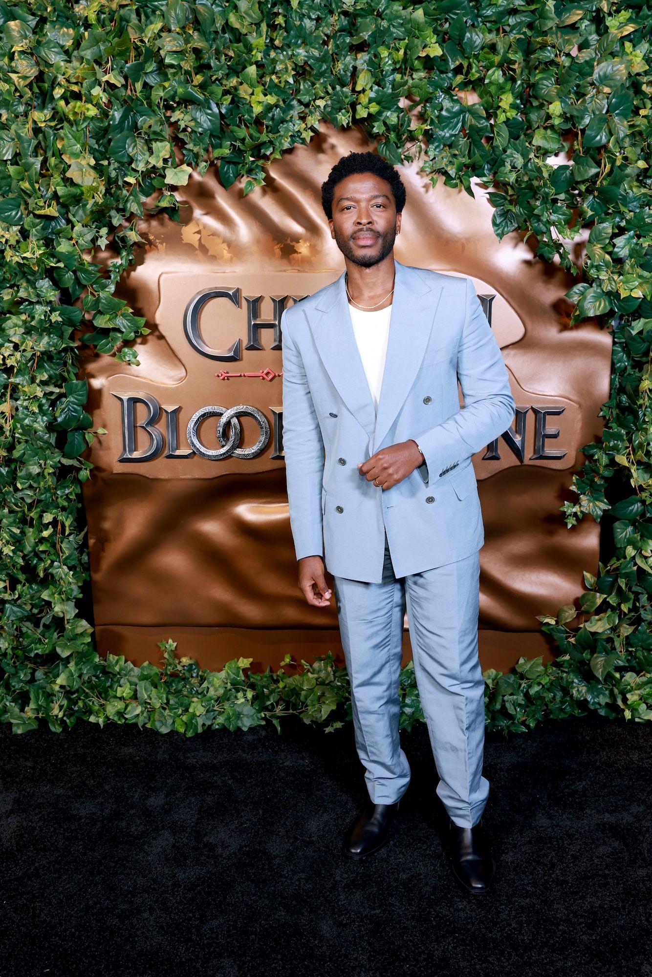 A man in a light gray suit stands in front of a large golden "Children of Blood and Bone" logo surrounded by greenery.