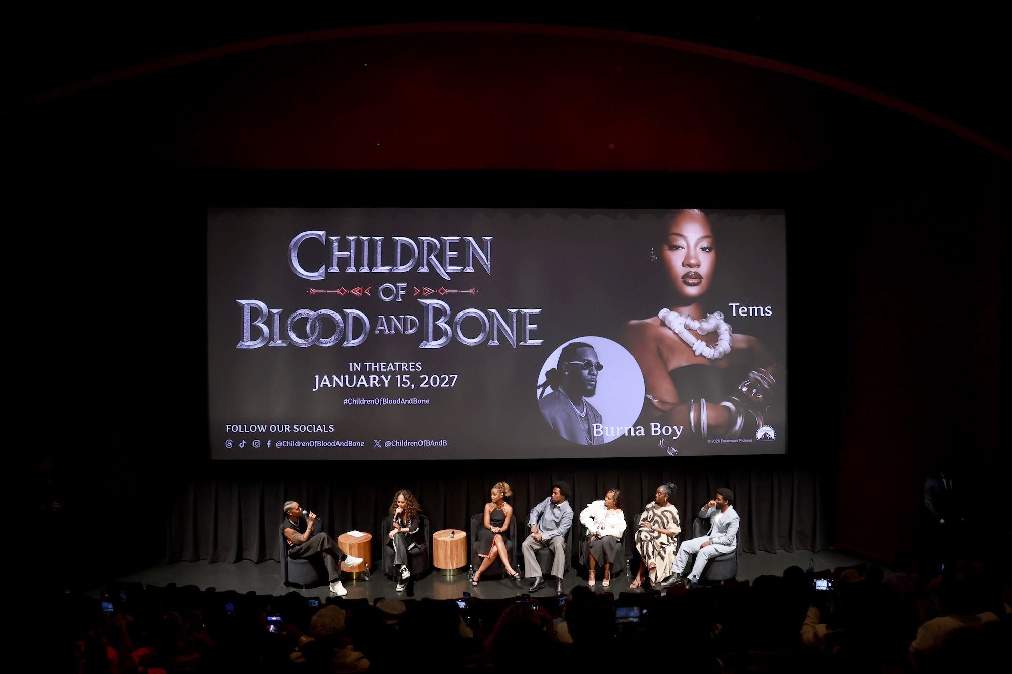 A group of people, including Lena Waithe, Gina Prince-Bythewood, Thuso Mbedu, Tosin Cole, Regina King, Lashana Lynch, and Zackary Momoh, speaking on stage in front of a large movie poster for "Children of Blood and Bone".
