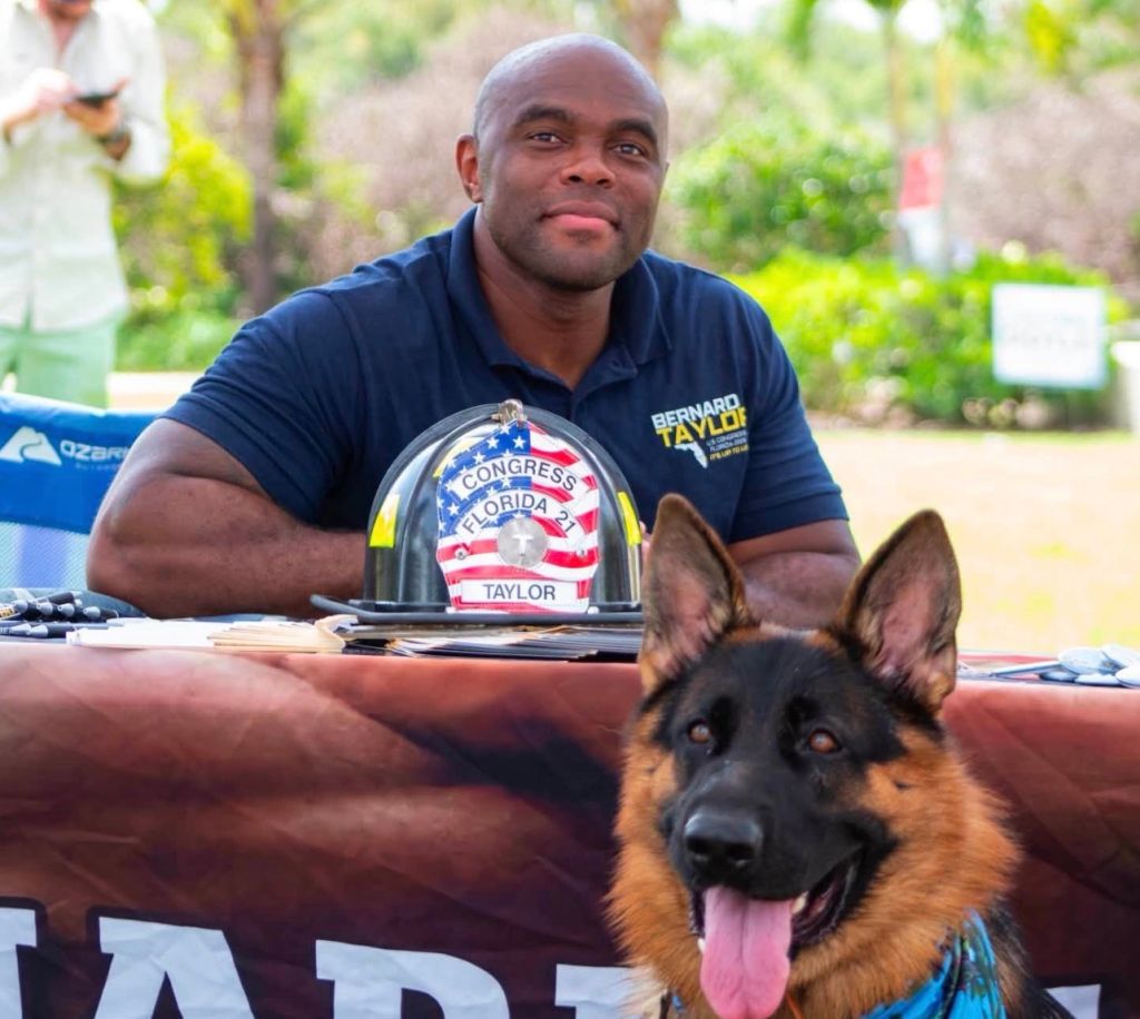 A man in a navy blue uniform with "Bernard Taylor" and "Congress Florida" on his helmet, sitting at a table with a German shepherd dog.