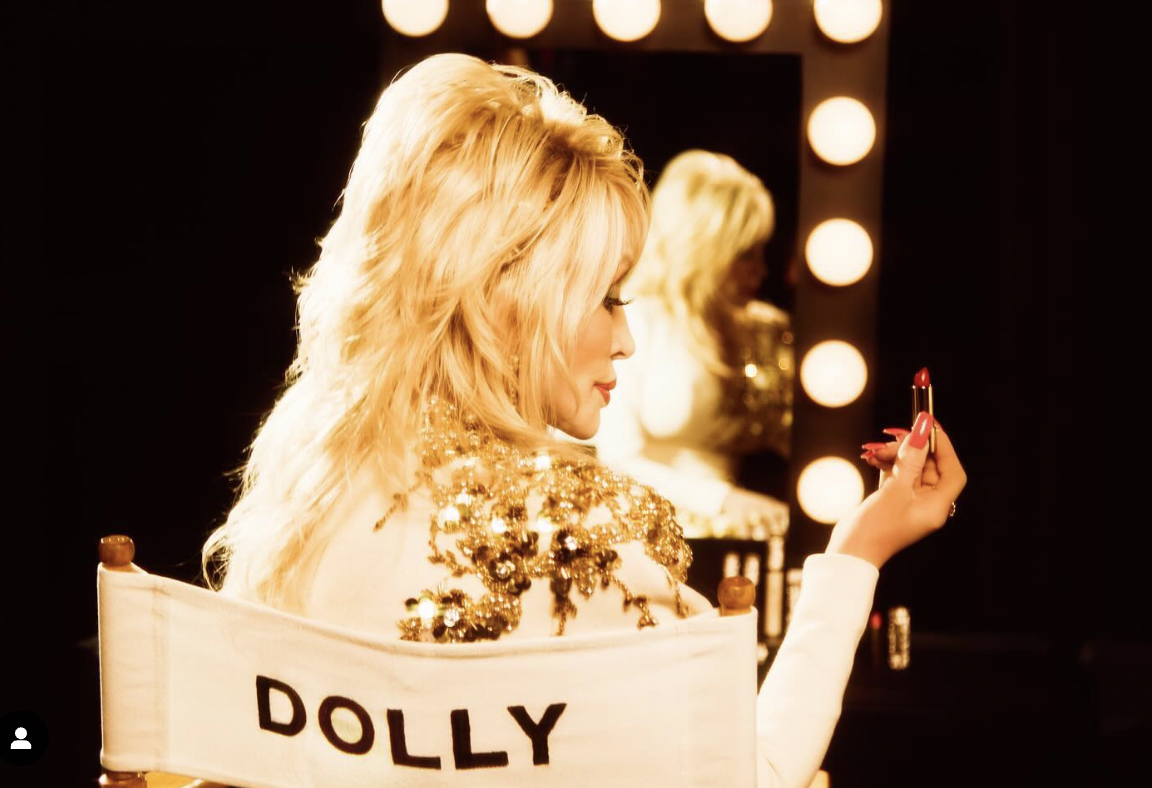 A woman with long, blonde hair applying lipstick in front of a mirror with bright lights around it, wearing a gold, sequined dress.
