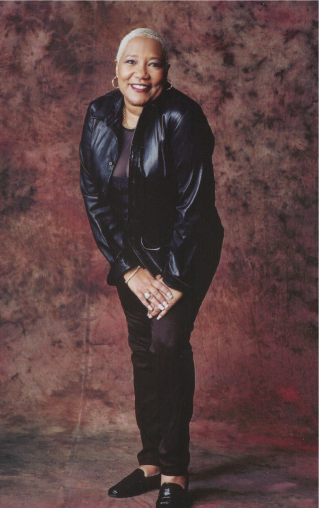 A smiling older woman wearing a black leather jacket stands against a maroon background.