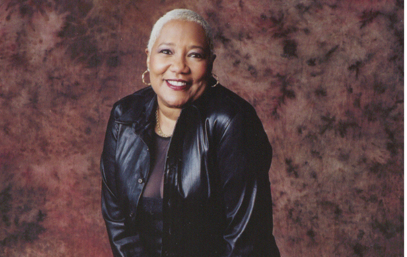 A smiling older woman wearing a black leather jacket stands against a maroon background.