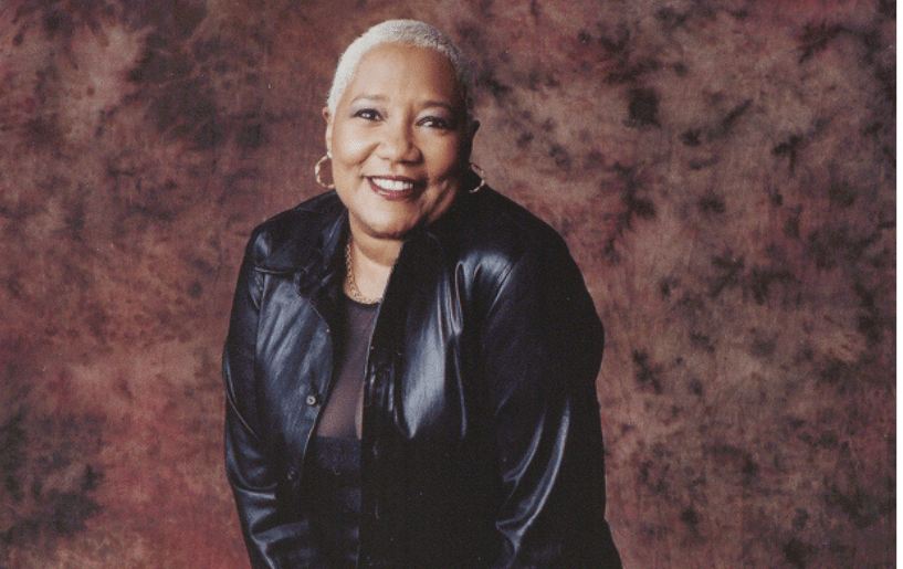 A smiling older woman wearing a black leather jacket stands against a maroon background.