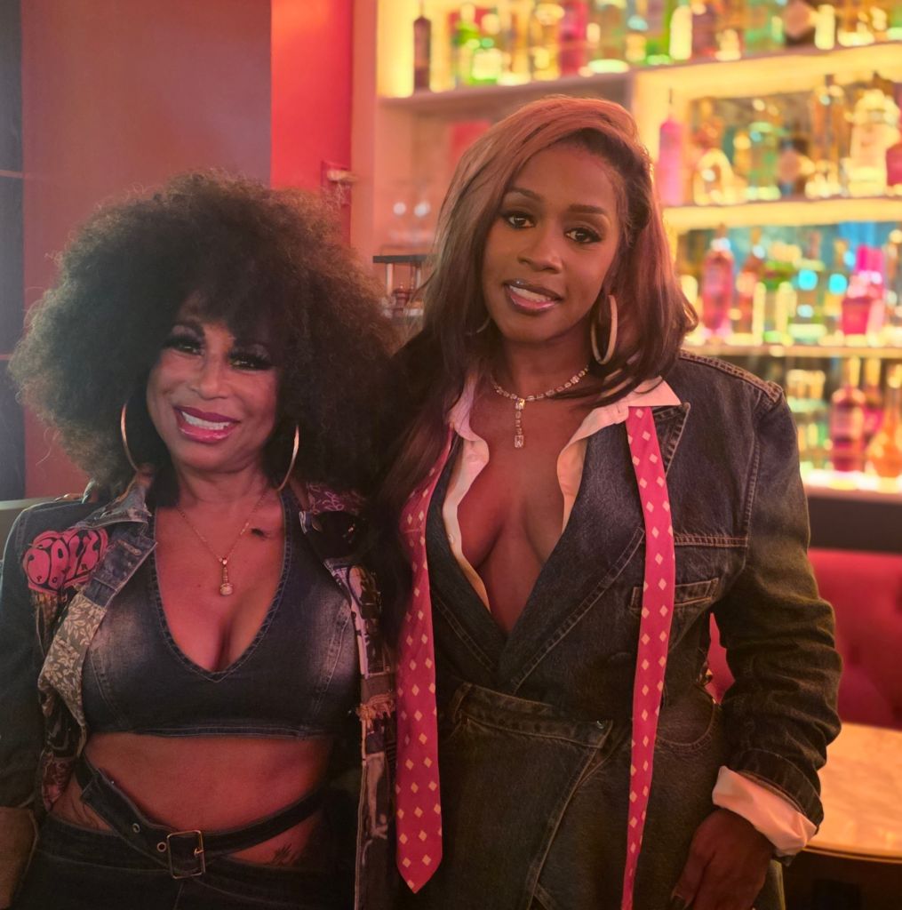 Two women in casual clothing posing together in a colorful, cluttered room with shelves of decorative items.