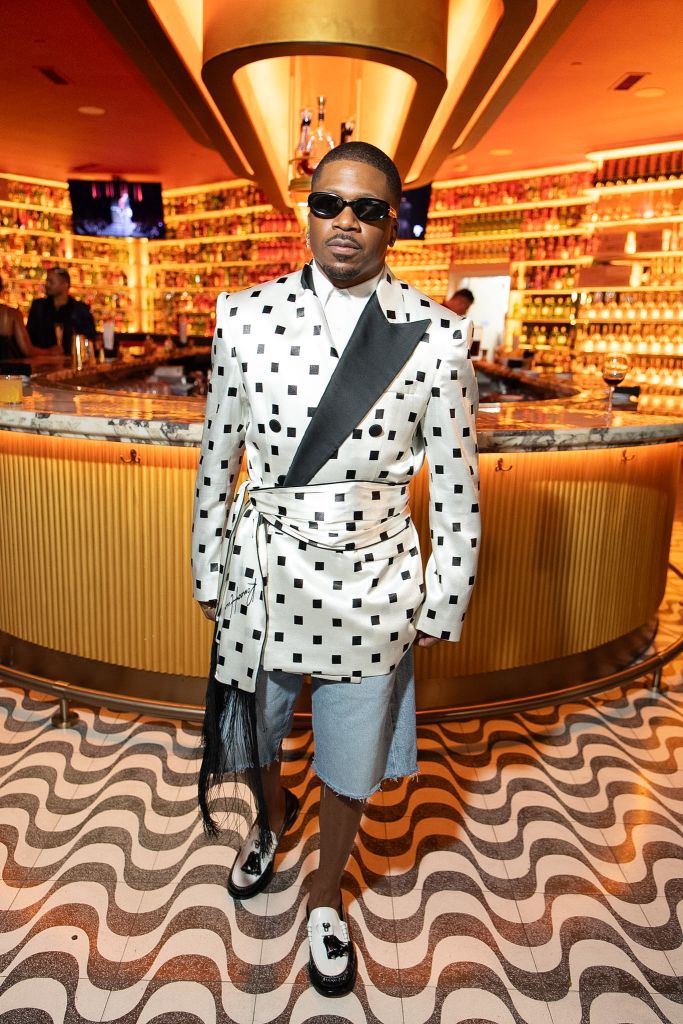 A well-dressed man wearing a black and white patterned suit and sunglasses stands in a lavishly decorated bar or lounge with a curved marble bar and ornate lighting fixtures.