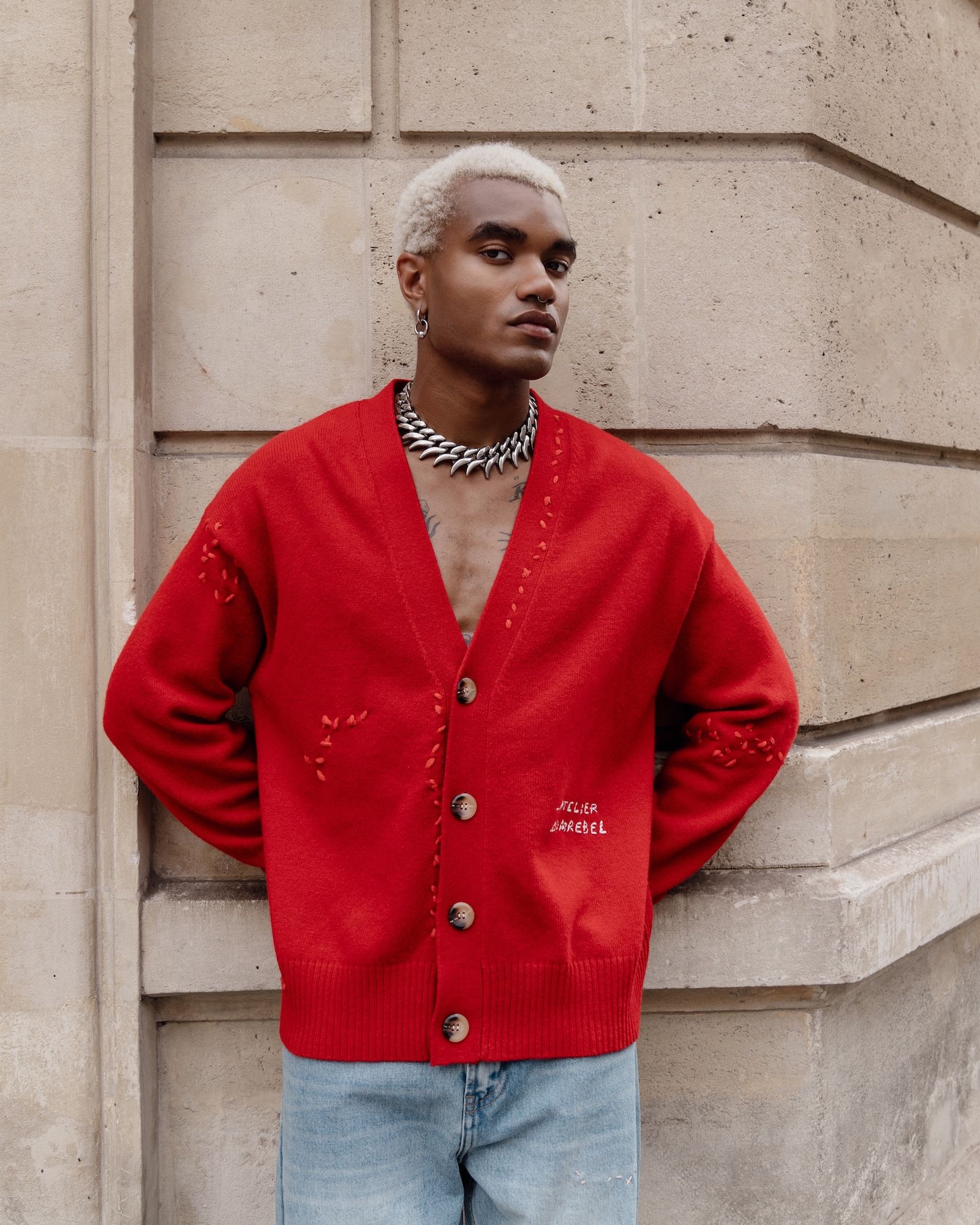 A person wearing a red cardigan with a silver necklace stands in front of a concrete wall.