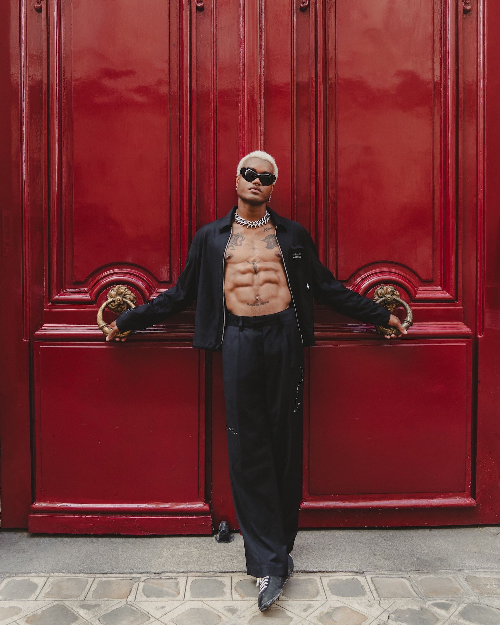 A person with a muscular, tattooed torso wearing sunglasses and a black jacket, standing in front of a vibrant red door.