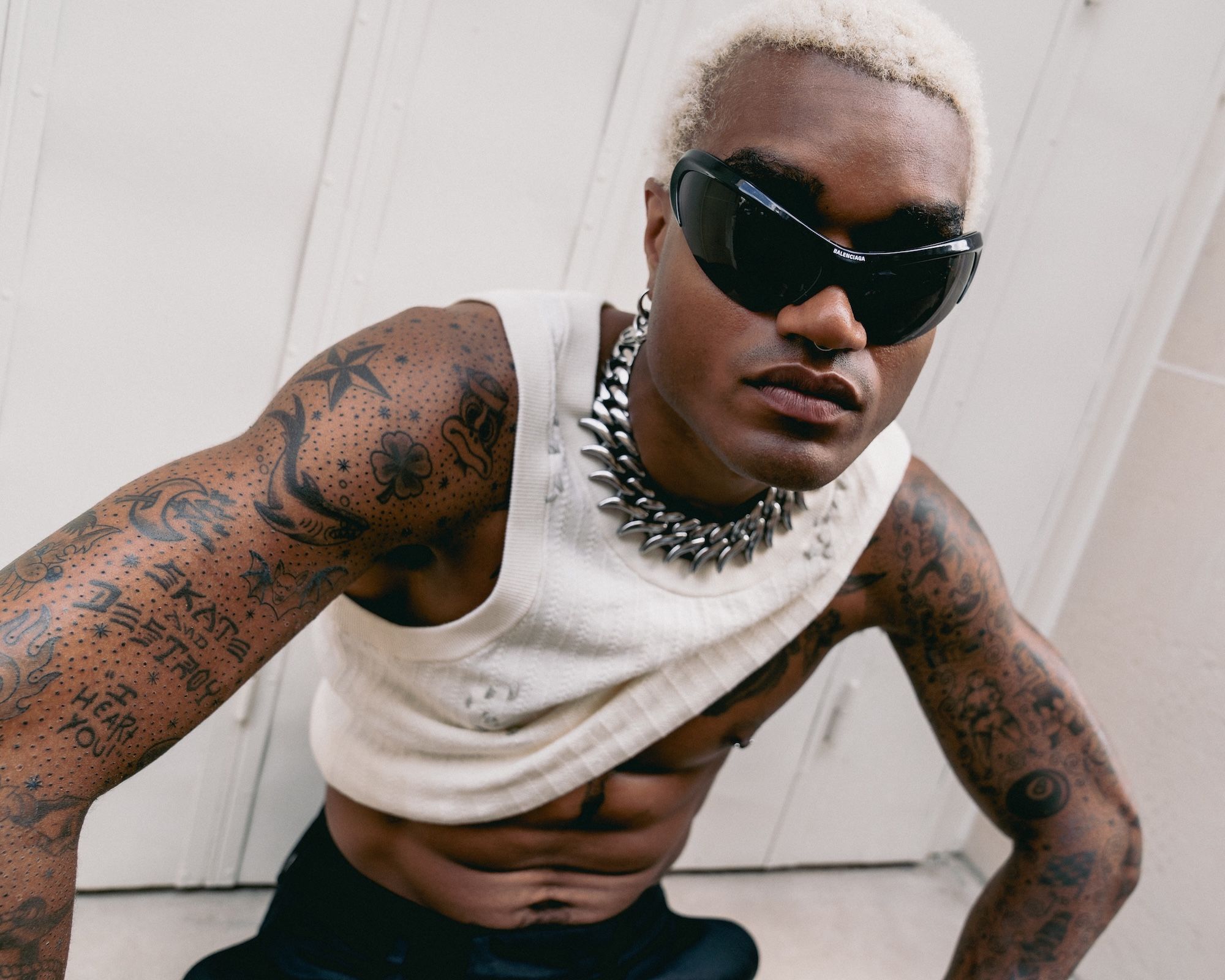Tattooed person wearing sunglasses and a necklace with a white tank top against a white wall.