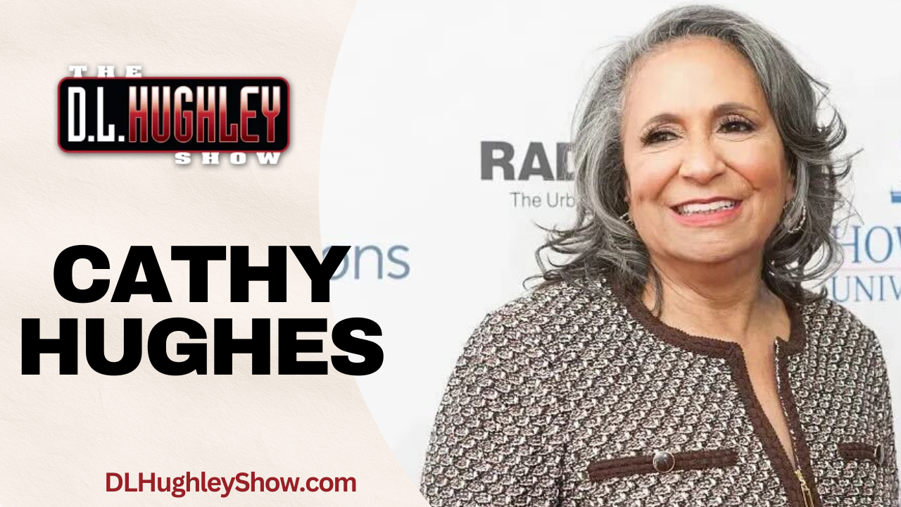 A smiling woman with gray hair wearing a patterned jacket, standing in front of a backdrop with the text "The D.L. Hughley Show" and "Cathy Hughes".